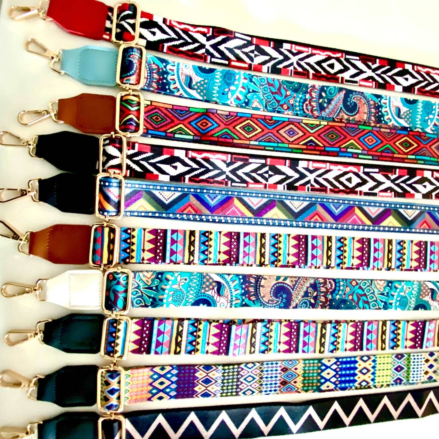 The image showcases a collection of Luxe Bohemian Guitar Style Shoulder Straps laid out horizontally. Each strap is adorned with vibrant, intricate patterns, featuring a mix of geometric shapes and vivid colors that exude a bohemian aesthetic. The straps are finished with sturdy hardware, suggesting a functional yet fashionable accessory designed to complement and enhance a variety of handbags or travel items. The overall presentation is rich and colorful, aligned with a luxurious and eclectic style