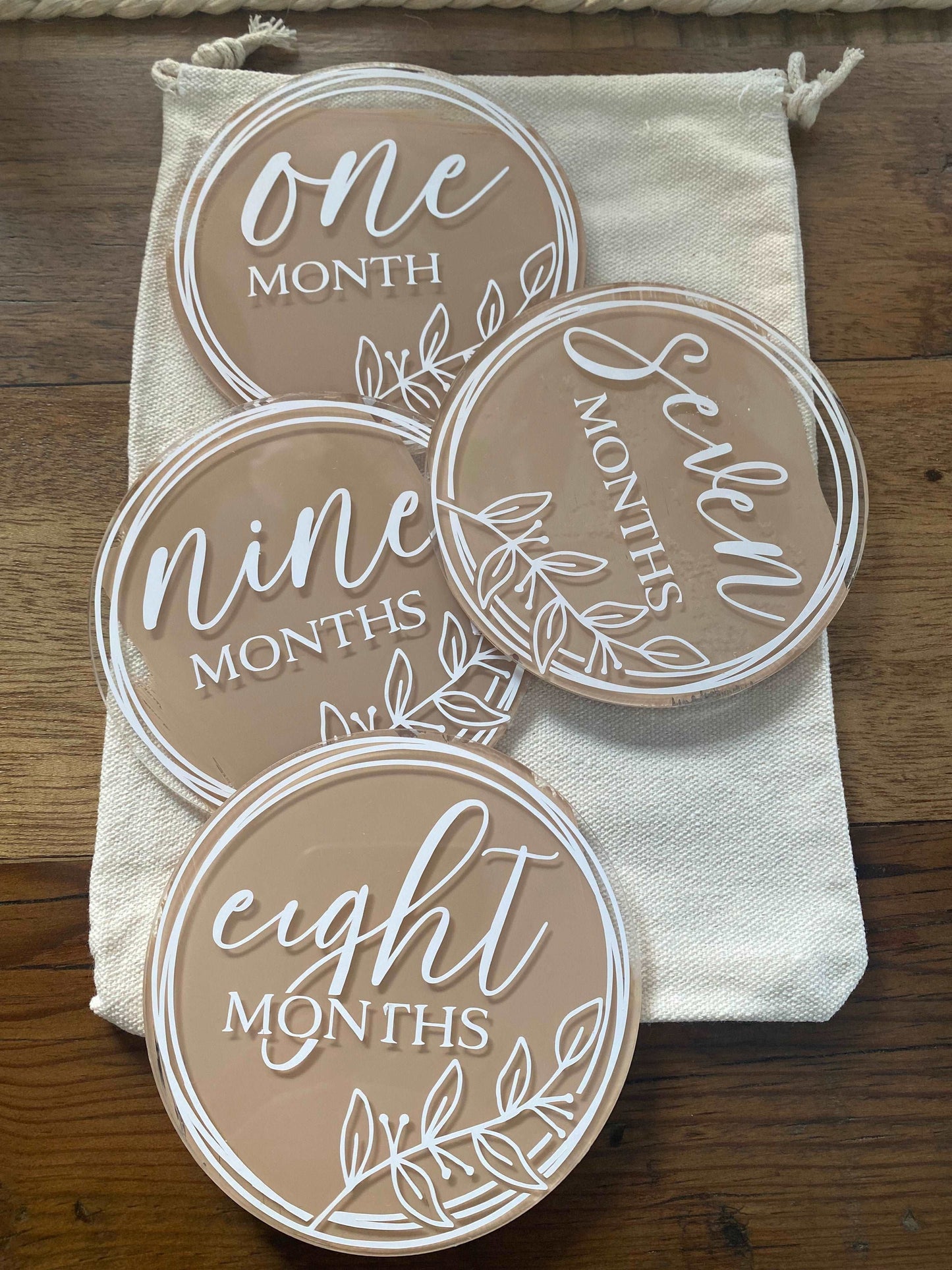 The image displays a set of round, flat discs, each with a different color background and a numeral indicating a month, from "one month" to "nine months," written in a stylish cursive font. These appear to be milestone markers used for photographing and celebrating a baby's monthly growth. Accompanying these discs are a pair of small, white baby shoes and a wooden bead garland, all arranged on a textured mat, suggesting a warm, homely atmosphere typical for a baby’s nursery.