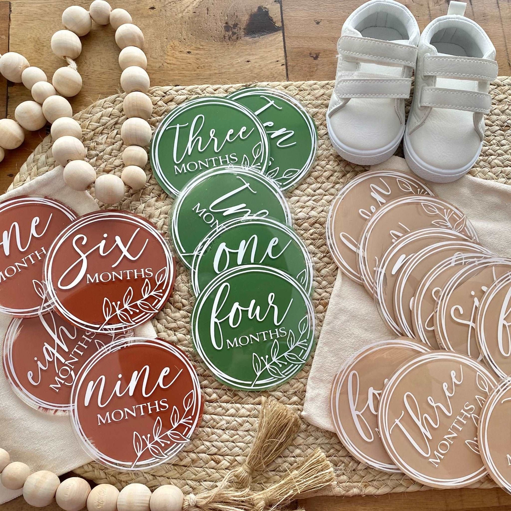 The image displays a set of round, flat discs, each with a different color background and a numeral indicating a month, from "one month" to "nine months," written in a stylish cursive font. These appear to be milestone markers used for photographing and celebrating a baby's monthly growth. Accompanying these discs are a pair of small, white baby shoes and a wooden bead garland, all arranged on a textured mat, suggesting a warm, homely atmosphere typical for a baby’s nursery.