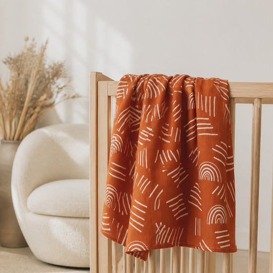 PJ Luxe terracotta baby muslin blanket with white rainbow print in a neutral nursery